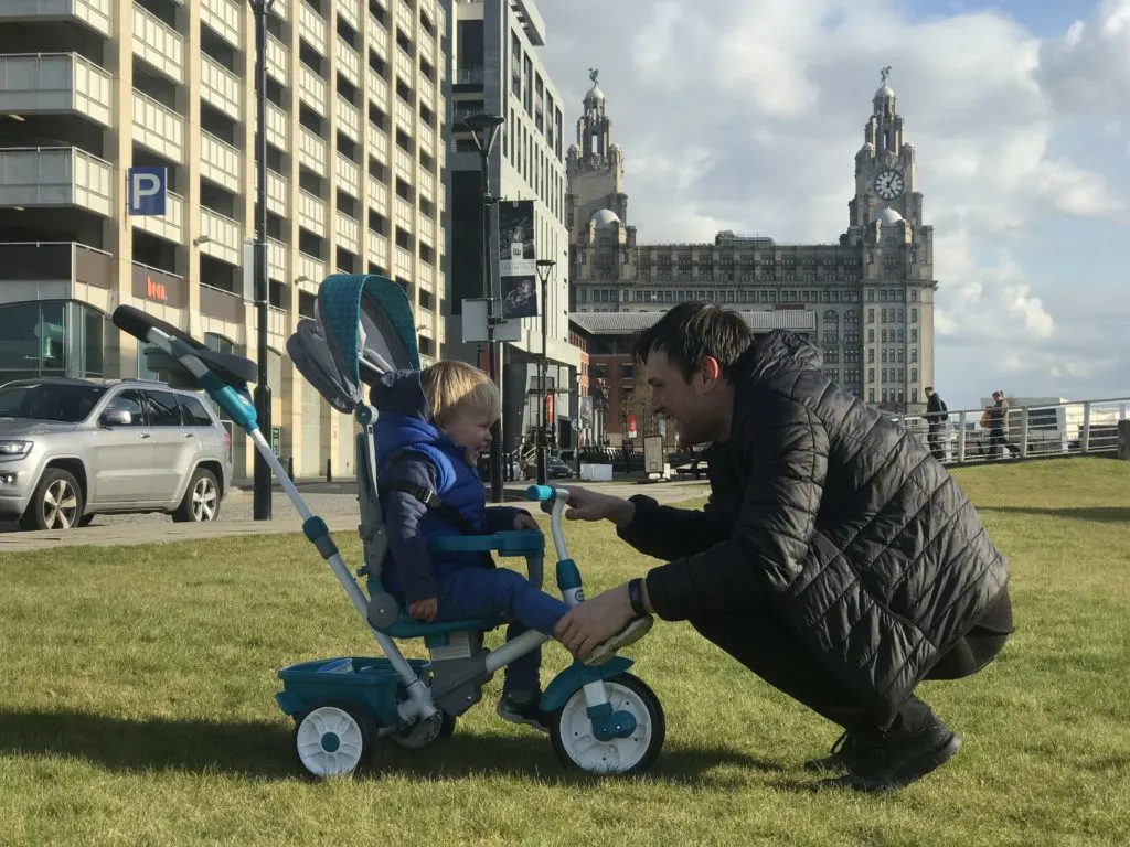 Dexter on his little tikes 4 in 1 trike infront of the liver building looking at his dad who is kneeling in front of him