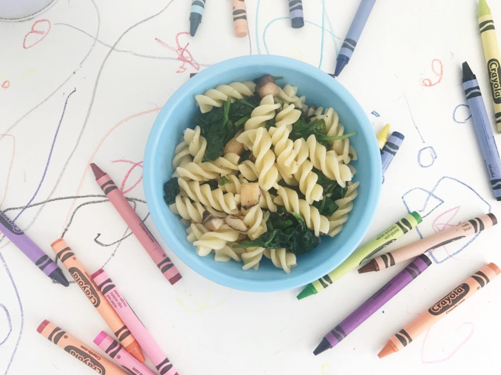 a bowl of mushroom and spinach toddler pasta served in a blue bowl on a background of drawn on paper and crayons scattered around