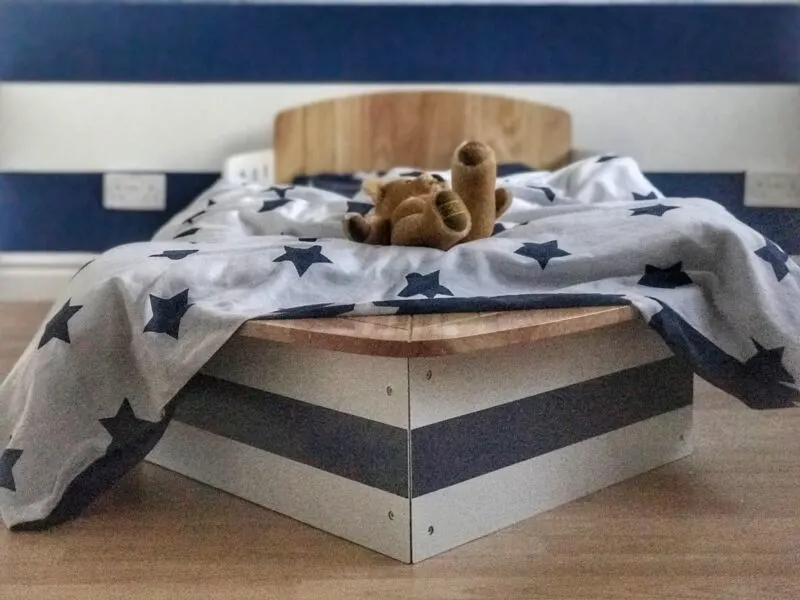 Dexter's toddler bed which is shaped like a boat with a white duvet with navy blue stars and a teddy bear placed on top