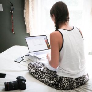 Woman sat on her bed using her laptop with her camera next to her