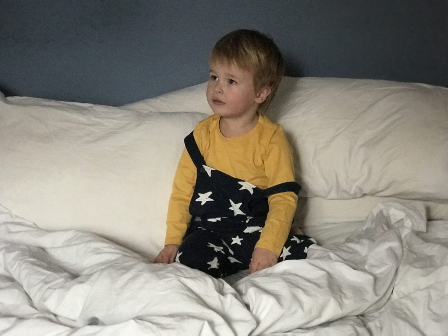 Dexter sitting on the bed wearing a yellow t shirt and navy blue dungarees with white stars on